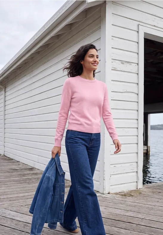 Femme en pull rose et jean tenant une veste en denim sur une passerelle près d’un hangar à bateaux.