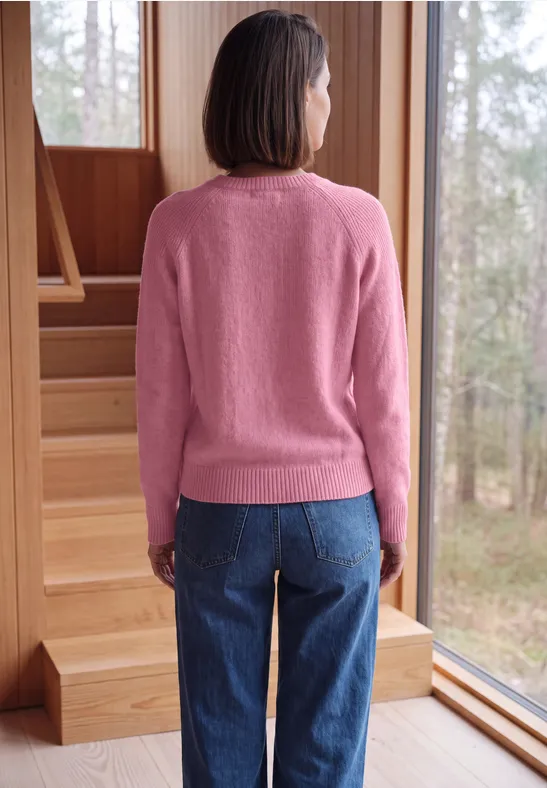 Personne de dos en pull rose et jean, debout près de grandes fenêtres en bois, regardant la forêt.