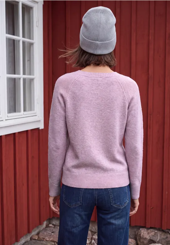 Personne en pull rose, bonnet gris et jean, de dos devant un mur en bois rouge.