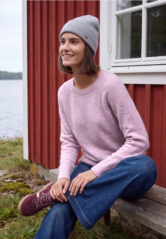 Femme souriante en pull rose, jean et bonnet gris assise sur une marche en bois devant une maison rouge au bord du lac.