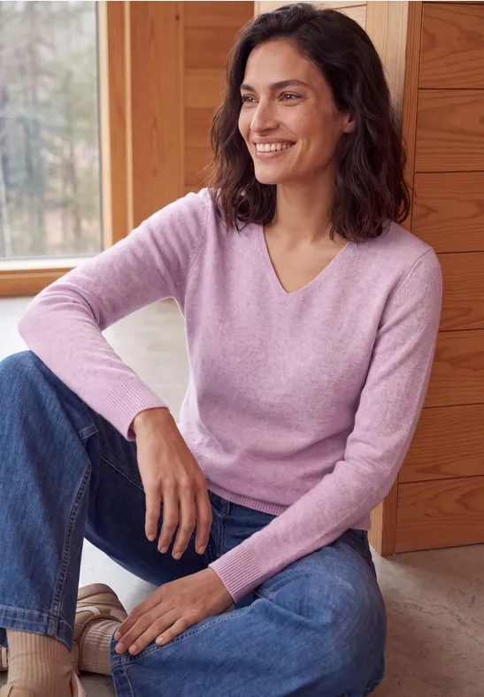 Une femme souriante en jean et pull violet pâle, assise devant un mur en bois.