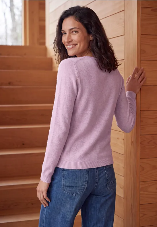 Une femme souriante en pull rose et jean se tient sur un escalier en bois tournant, regard par-dessus l'épaule.