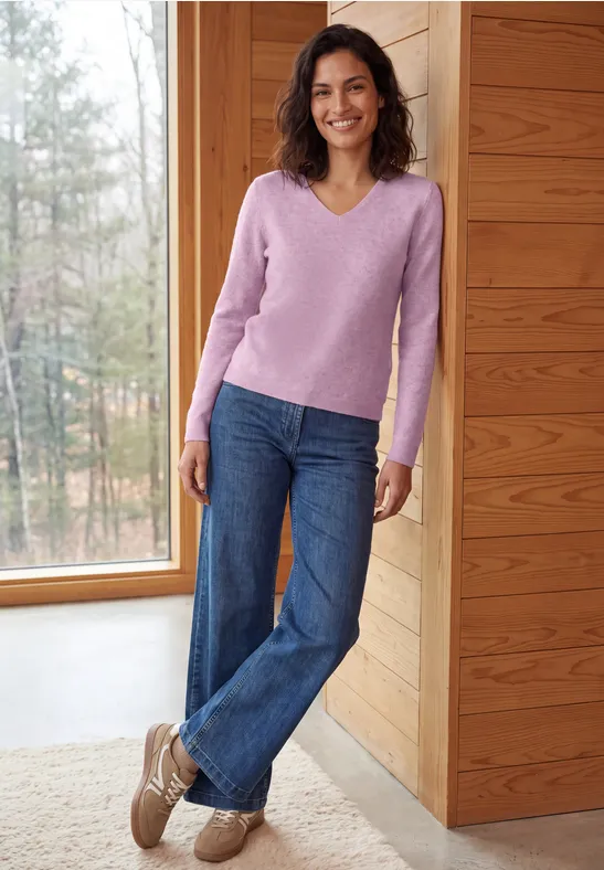 Une femme en pull rose et jean sourit, devant une baie vitrée en bois avec vue sur la nature.