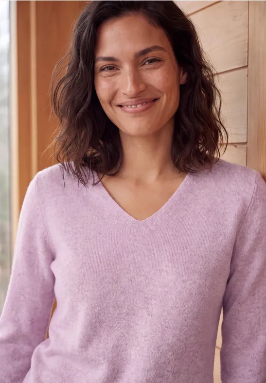 Une femme souriante en pull lilas appuyée contre un mur en bois.