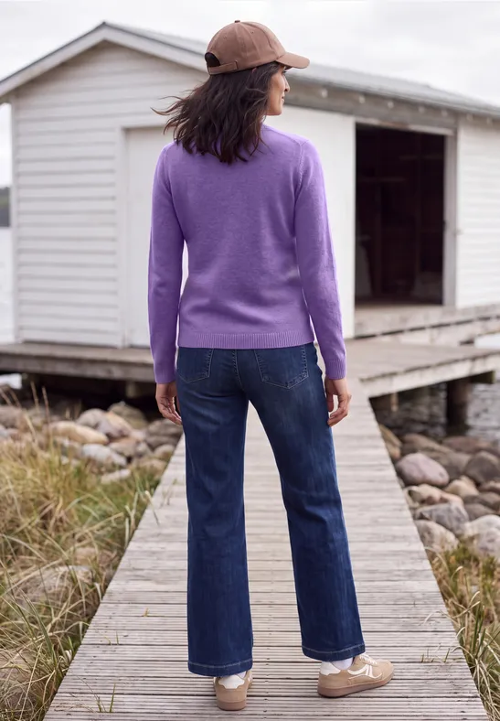 Femme en pull violet, jean et casquette se tient sur une passerelle en bois devant un petit chalet blanc.