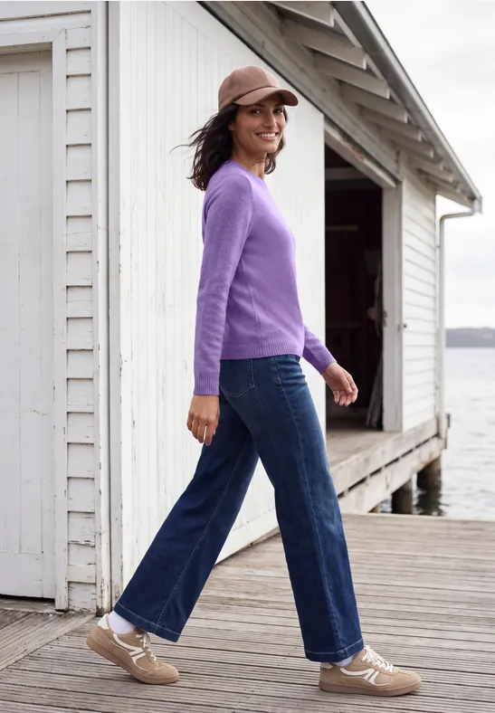 Femme en pull violet, jean et casquette, souriante, marchant sur une jetée en bois au bord de l'eau.