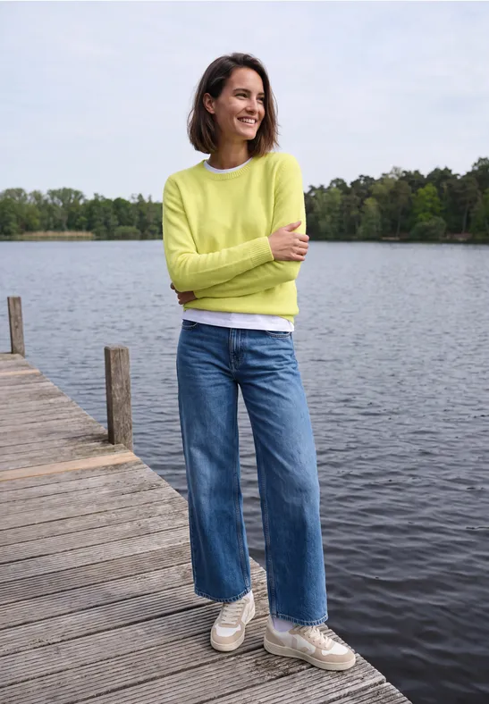 Femme souriante sur une jetée au bord d'un lac, vêtue d'un pull vert clair, jean et baskets blanches.