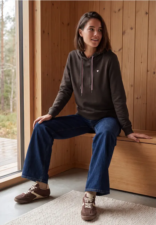 Femme en sweat à capuche marron et jean bleu assise confortablement sur un banc en bois près d'une fenêtre.