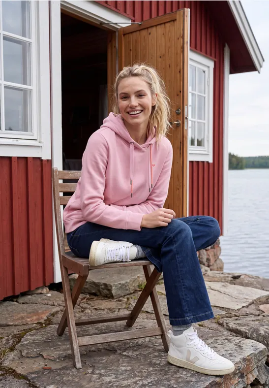 Femme en sweat à capuche rose assise sur une chaise en bois devant un chalet rouge au bord du lac.
