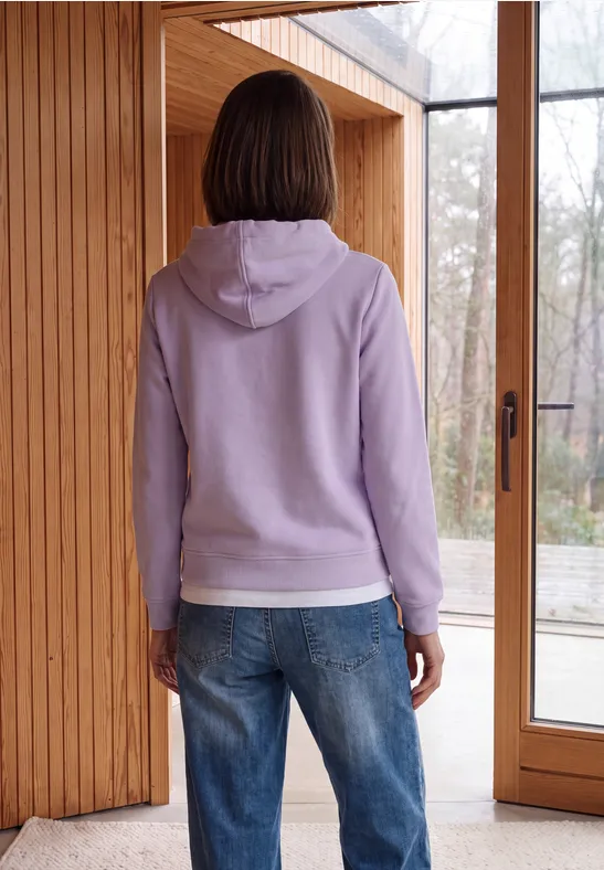 Une personne en sweat à capuche violet et jean se tient devant une grande porte-fenêtre en bois.