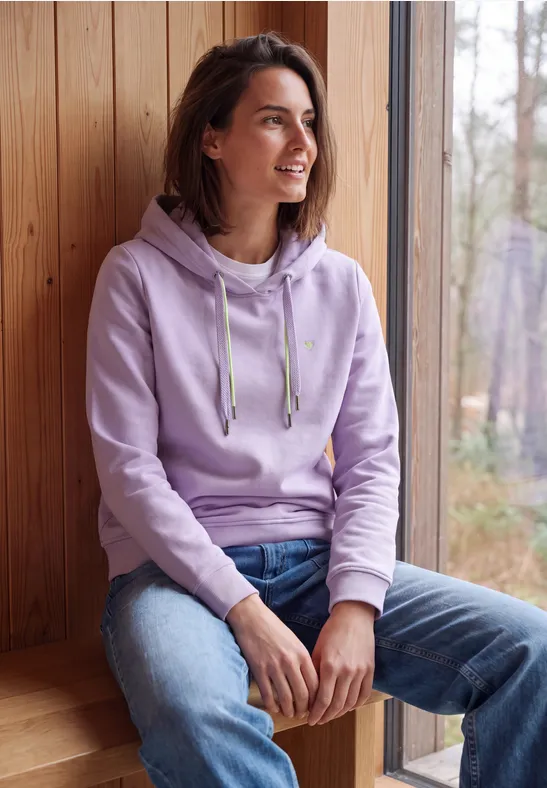 Jeune femme en sweat à capuche violet assise près d'une fenêtre en bois, souriant et regardant dehors.