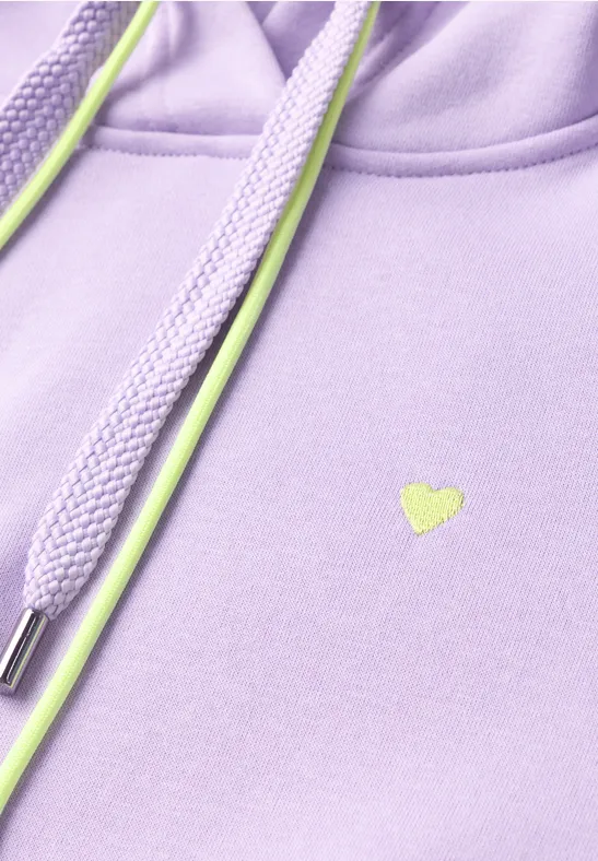 Gros plan d'un sweat à capuche lavande avec cordons vert néon et petit logo cœur.