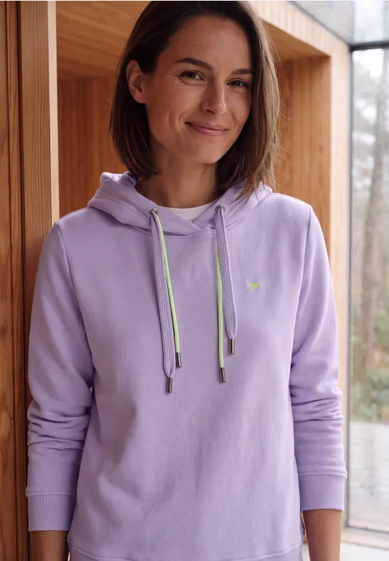 Femme souriante aux cheveux courts, portant un sweat à capuche violet devant une baie vitrée.