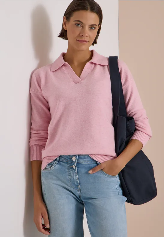 Une jeune femme porte un pull polo rose, un jean et un sac bleu marine à l'épaule.