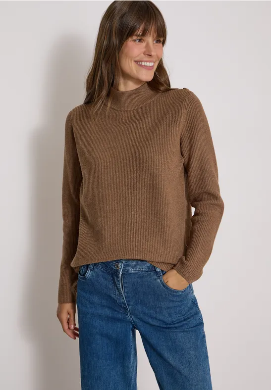 Eine lächelnde Frau trägt einen braunen Pullover und blaue Jeans vor hellem Hintergrund.
