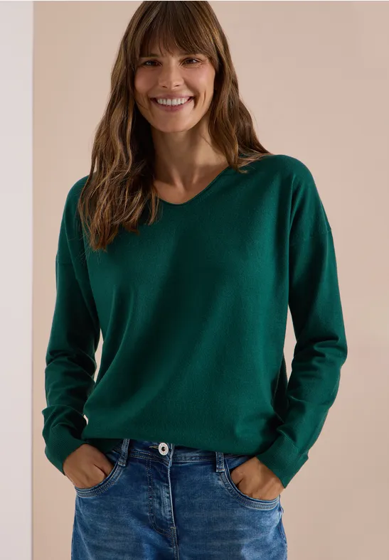 Une femme souriante portant un pull vert et un jean devant un fond neutre.