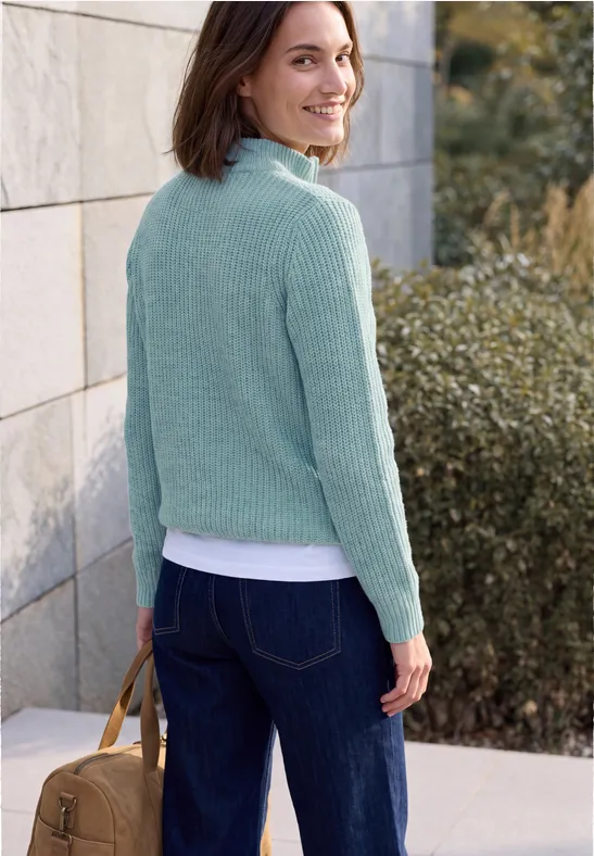 Femme en pull vert menthe et jean, souriante avec un sac marron, debout devant un mur et des buissons.