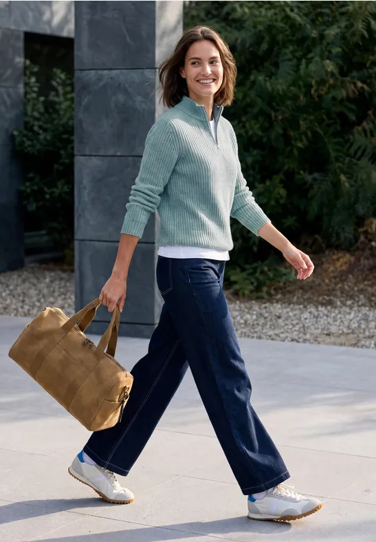 Femme souriante marchant dehors, porte un pull bleu clair, un pantalon bleu et un sac en daim marron.