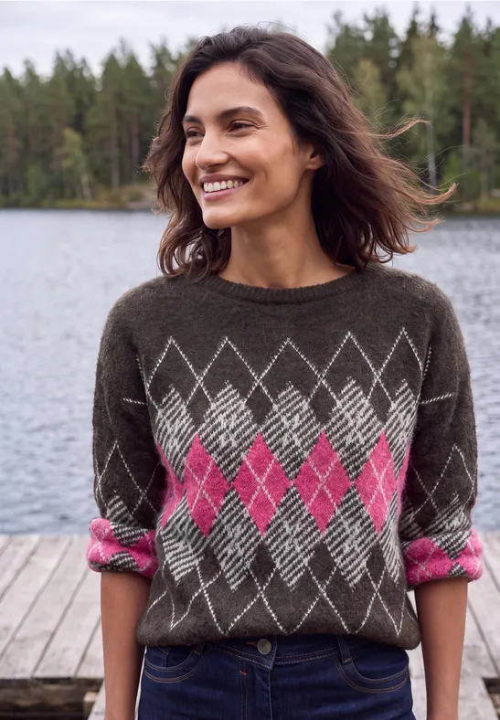Een glimlachende vrouw in een bruine trui met roze ruitmotief, staand op een steiger voor een meer in het bos.