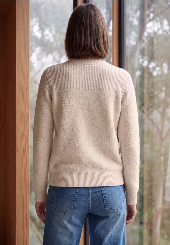 Frau von hinten in hellem Pullover und Jeans, steht vor großem Fenster mit Blick auf Bäume.