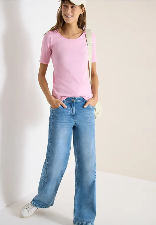 Femme en t-shirt rose, jean large et baskets blanches devant un mur blanc.