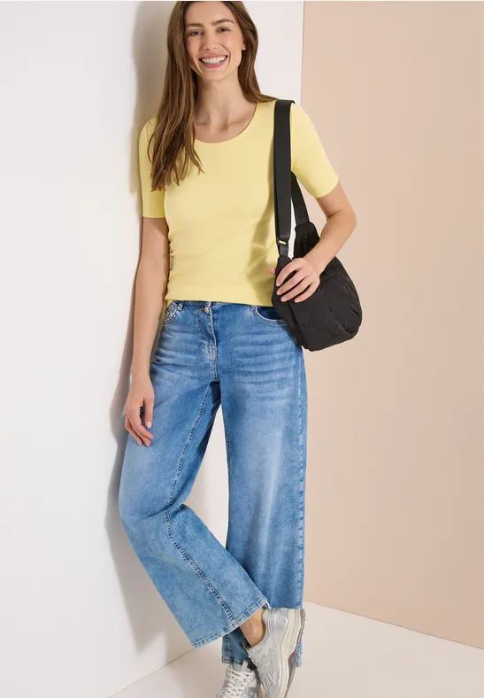 Frau in gelbem T-Shirt, blauen Jeans und Tasche steht vor hellem Hintergrund.