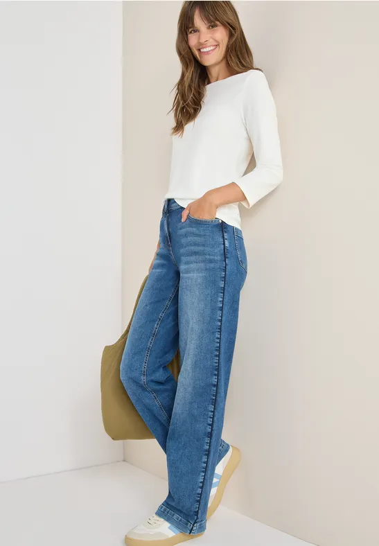 Frau mit weißem Oberteil, blauer Jeans und Sneakers lächelt an der Wand.