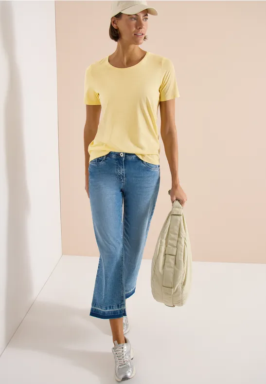 Frau in gelbem T-Shirt, Jeans und Cap trägt Tasche und Sneakers.