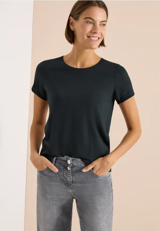 Frau in schwarzem T-Shirt und grauer Jeans, steht mit Händen in den Taschen vor hellem Hintergrund.