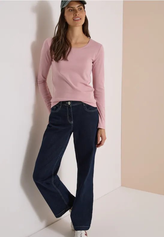 Une femme appuyée contre un mur porte un top rose, un jean large et une casquette.