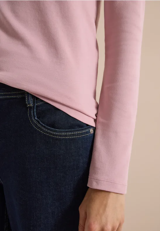 Gros plan sur une personne en haut rose manches longues et jean sombre, main le long du corps.