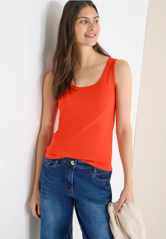 Frau in rotem Tanktop und Jeans lehnt an weißer Wand und hält eine beige Jacke.