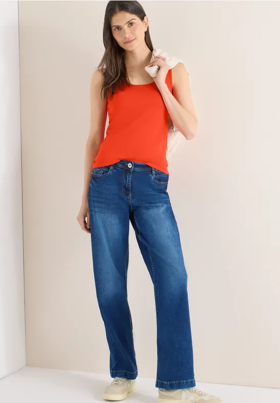 Frau in rotem Top und blauen Jeans steht locker an einer weißen Wand.
