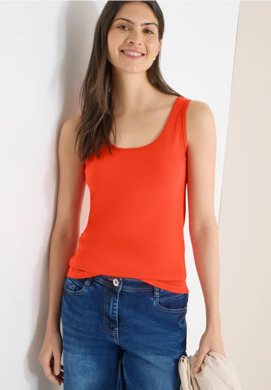 Frau in orangefarbenem ärmellosen Top und blauer Jeans steht vor hellem Hintergrund.
