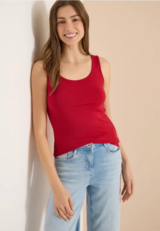 Eine Frau lehnt lässig an einer Wand und trägt ein rotes Tanktop und helle Jeans.