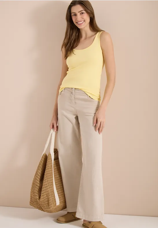 Femme en débardeur jaune et pantalon large beige, tenant un sac en osier devant un fond clair.