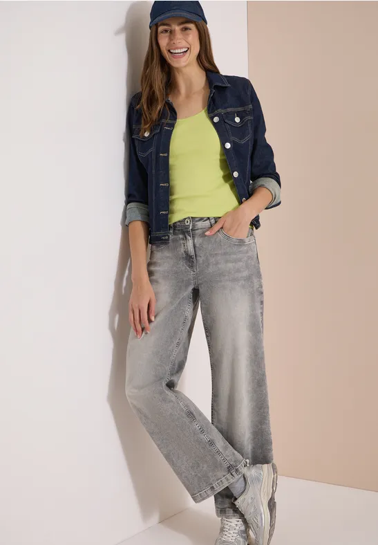 Frau lehnt an Wand, trägt Jeansjacke, grünes Top, weite graue Jeans und weiße Sneaker.