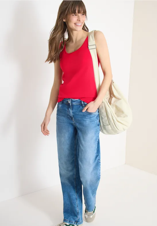 Frau trägt rotes Top, weite Jeans und Sandalen, mit einer großen hellen Tasche an der Schulter.