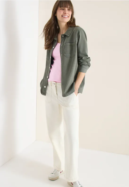 Femme en surchemise verte, débardeur rose, pantalon blanc et baskets dans un cadre épuré.