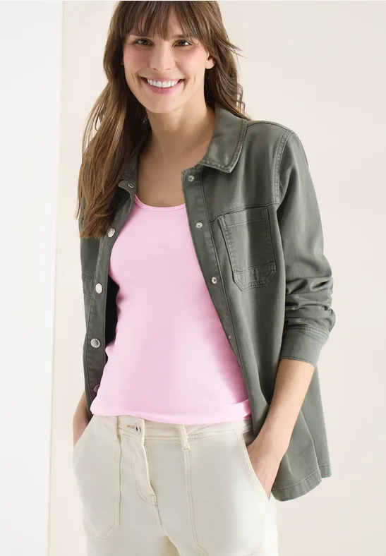 Femme portant un débardeur rose, une surchemise kaki et un pantalon clair, mains en poches.