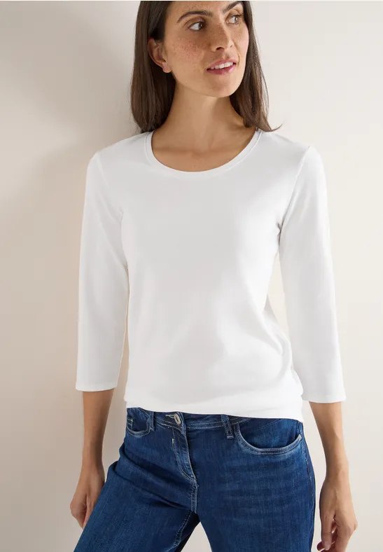 Frau mit weißem Langarmshirt und blauer Jeans steht vor hellem Hintergrund.