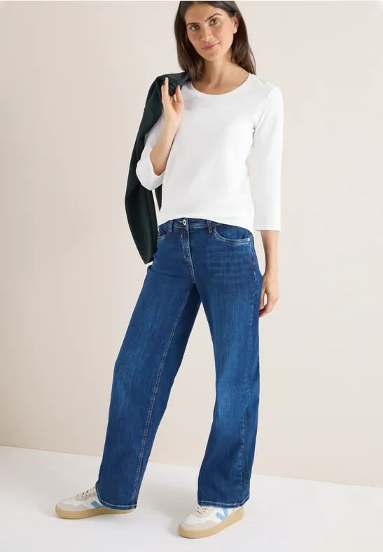Frau in weißem Shirt und weiter Jeans, Jacke über der Schulter.
