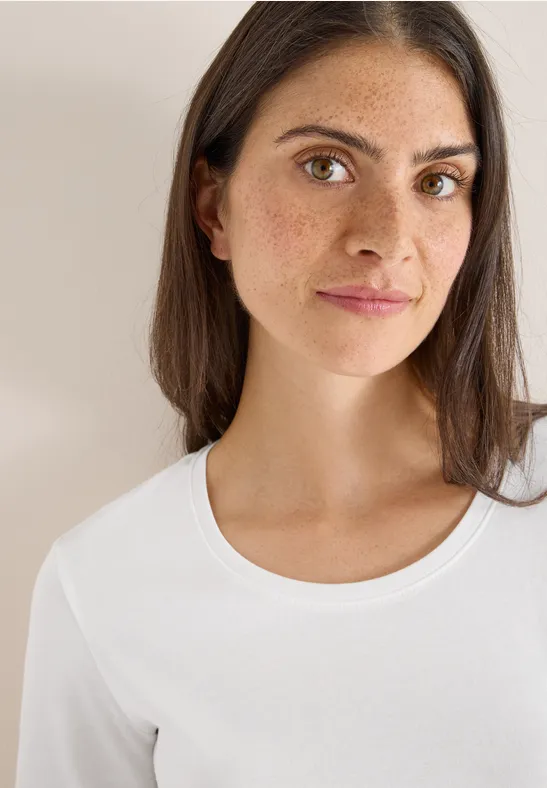 Frau mit Sommersprossen, langen braunen Haaren und weißem Shirt blickt in die Kamera.