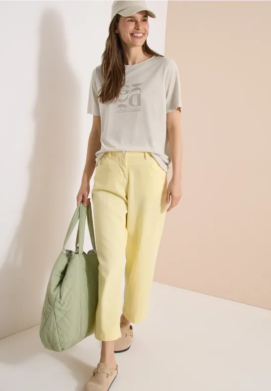 Frau in grauem T-Shirt, gelben Hosen und grüner Tasche vor hellem Hintergrund.