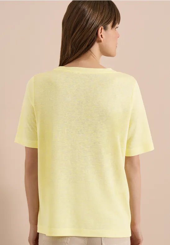 Frau trägt schlichtes gelbes T-Shirt, steht mit dem Rücken zur Kamera vor beigem Hintergrund.