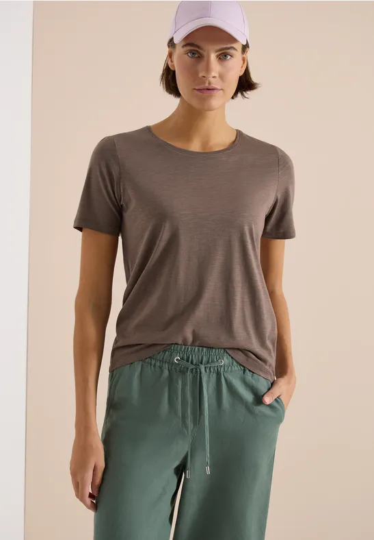 Frau in grauem T-Shirt und grüner Hose vor hellem Hintergrund.