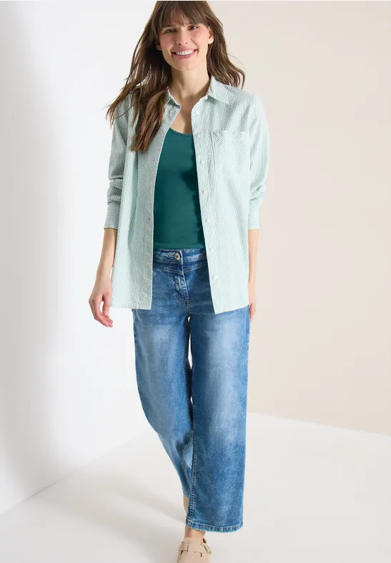 Frau trägt Jeans, grünes Top und helle, gestreifte Hemdbluse, steht vor neutralem Hintergrund.
