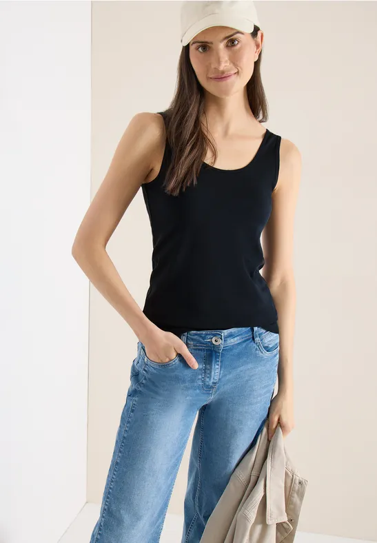 Frau trägt schwarzes Tanktop, blaue Jeans und hält eine beige Jacke in der Hand.