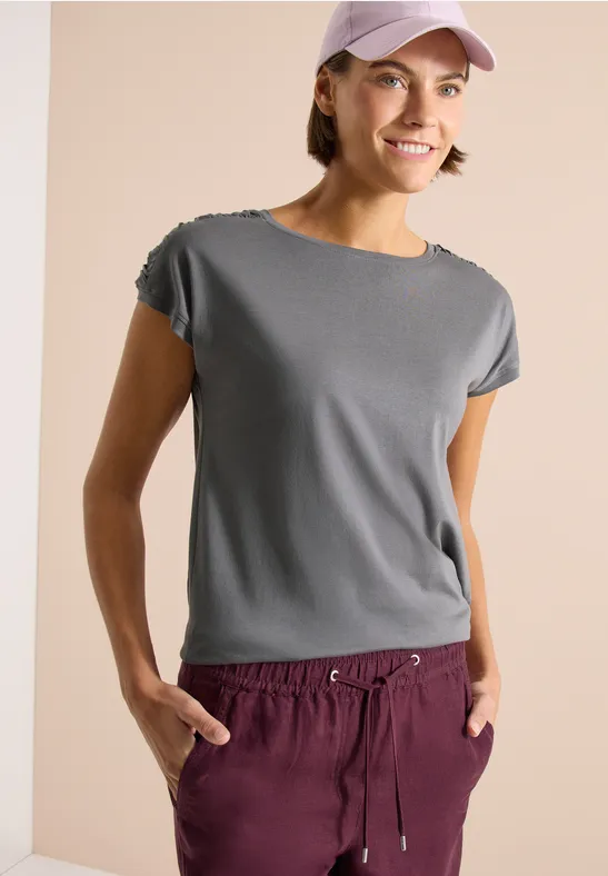 Frau trägt graues T-Shirt, weinrote Jogginghose und rosa Basecap vor neutralem Hintergrund.