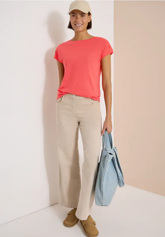 Person trägt korallenrotes T-Shirt, beige Hose und hellblaue Tasche vor neutralem Hintergrund.